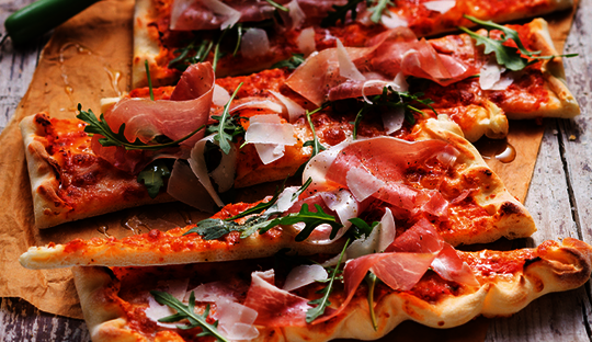 [Translate to Magyar:] Bild einer Italian Style Pizza, zubereitet mit Wewalka Pizzateig gerollt auf Backpapier, angerichtet auf einem Holzbrett und belegt mit Tomaten, Prosciutto und Rucola
