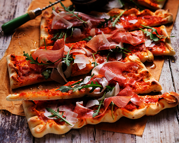 [Translate to Magyar:] Bild einer Italian Style Pizza, zubereitet mit Wewalka Pizzateig gerollt auf Backpapier, angerichtet auf einem Holzbrett und belegt mit Tomaten, Prosciutto und Rucola