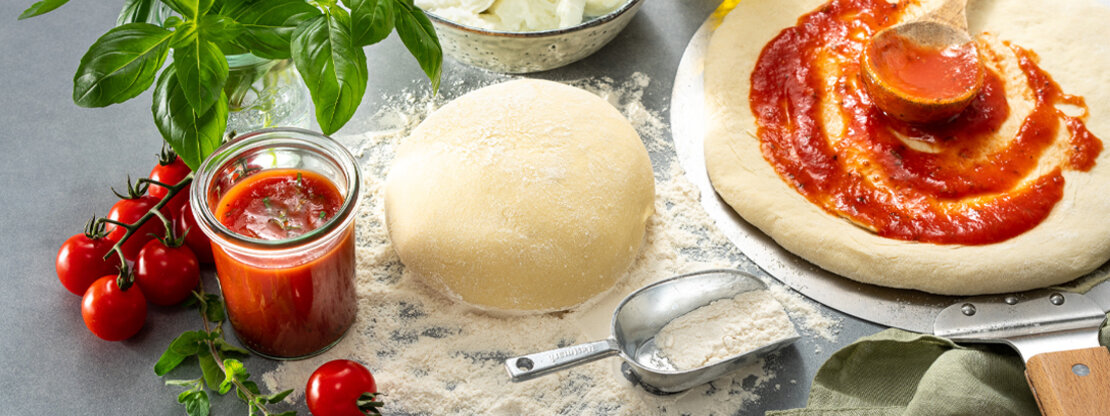 Picture of a Wewalka sourdough-ball on a kitchen workop, already with tomato sauce on the prepared pizza, decorated with basil and fresh tomatoes on the left side