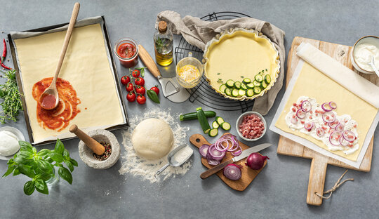 Picture of a variation of Wewalka fresh doughs, which are being prepared on a kitchen worktop with various hearty ingredients, including pizza crust pre-rolled on baking paper, a sourdough ball, short crust as a quiche and a flatbread dough