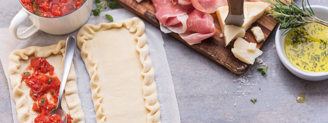 Picture of pizzettas, being prepared with Wewalka pizza crust pre-rolled on baking paper on a kitchen worktop, topped with tomato sauce, prosciutto ham, parmiggiano cheese and fresh olive oil