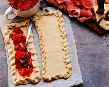 Picture of pizzettas, being prepared with Wewalka pizza crust pre-rolled on baking paper on a kitchen worktop, topped with tomato sauce, prosciutto ham, parmiggiano cheese and fresh olive oil